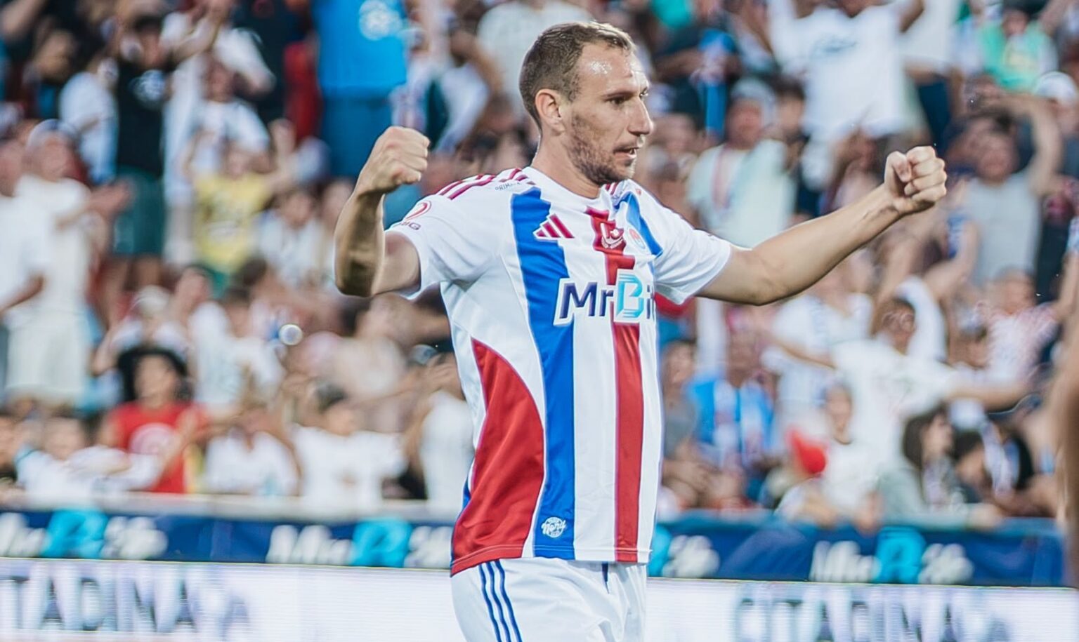Soccer player in white jersey with blue and red stripes celebrates with arms raised as crowd cheers behind him