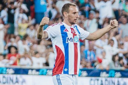 Soccer player in white jersey with blue and red stripes celebrates with arms raised as crowd cheers behind him