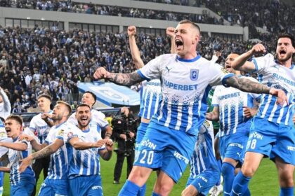 Group of football players in blue-and-white striped kits celebrate a goal on the field as fans cheer in the stands behind them.