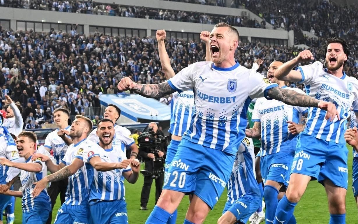 Group of football players in blue-and-white striped kits celebrate a goal on the field as fans cheer in the stands behind them.