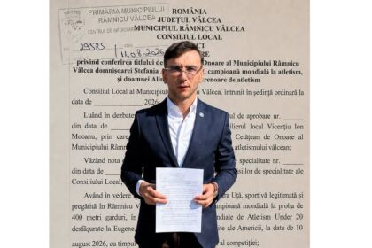 Man in a navy suit holding a white document in front of a large wall with Romanian text, likely at a municipal building.