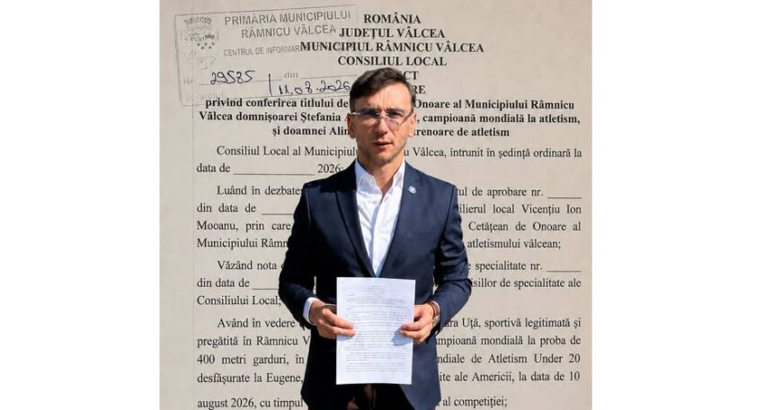 Man in a navy suit holding a white document in front of a large wall with Romanian text, likely at a municipal building.