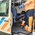 Paramedics in orange reflective uniforms assist a patient on a stretcher inside an ambulance, door open for loading.
