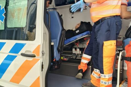 Paramedics in orange reflective uniforms assist a patient on a stretcher inside an ambulance, door open for loading.