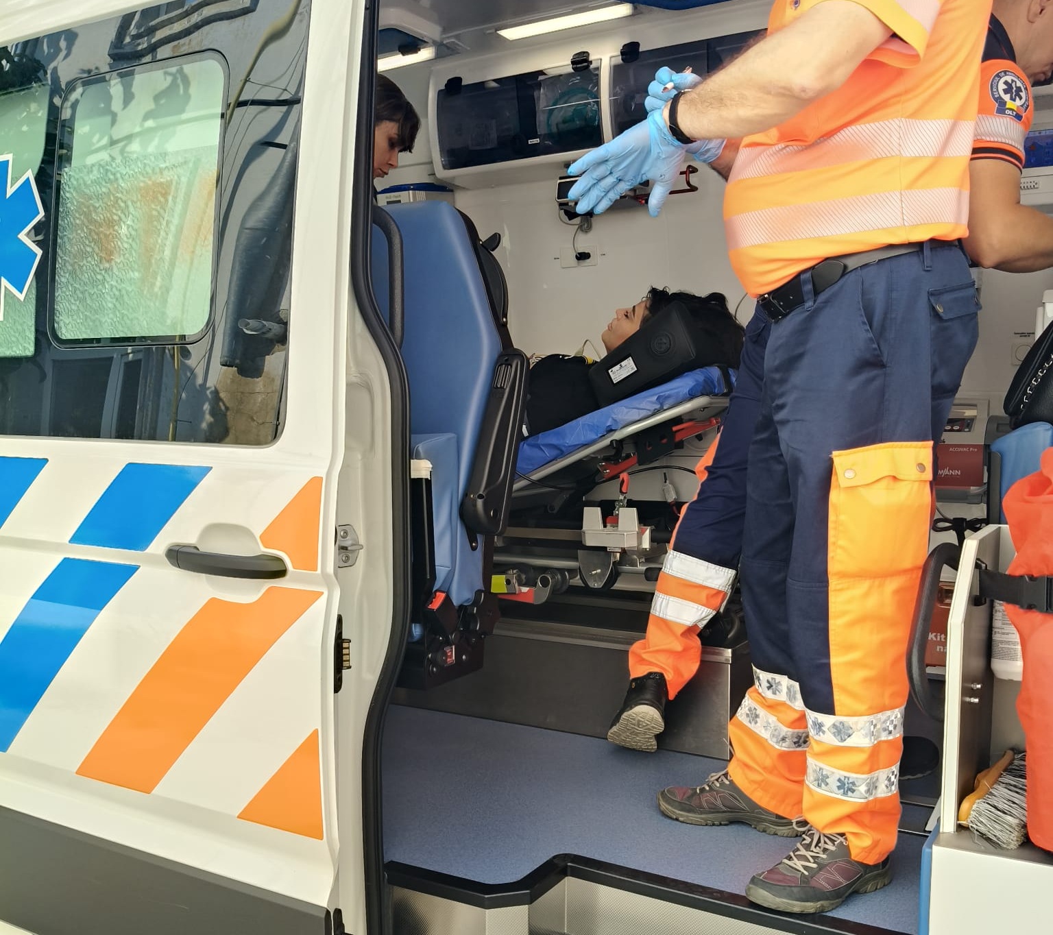 Paramedics in orange reflective uniforms assist a patient on a stretcher inside an ambulance, door open for loading.