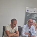 Two men sit at a conference table during a discussion, with a banner visible in the background.