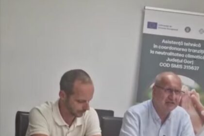 Two men sit at a conference table during a discussion, with a banner visible in the background.