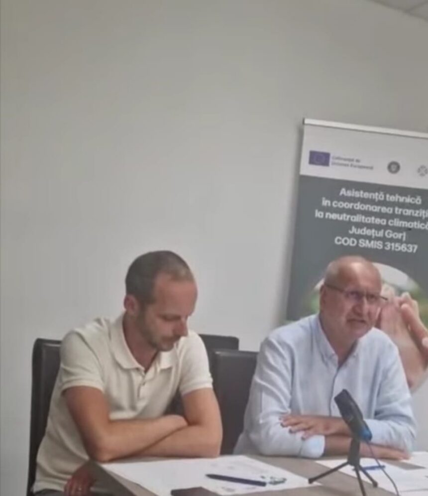 Two men sit at a conference table during a discussion, with a banner visible in the background.