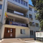 Facade of a white multi-story government building with stairs and a blue sign reading 'Inspectoratul Școlar Județean Gorj' above the entrance; Romanian flag nearby.