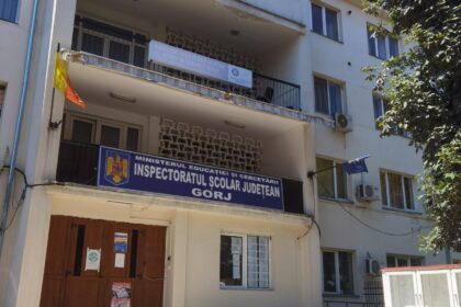 Facade of a white multi-story government building with stairs and a blue sign reading 'Inspectoratul Școlar Județean Gorj' above the entrance; Romanian flag nearby.