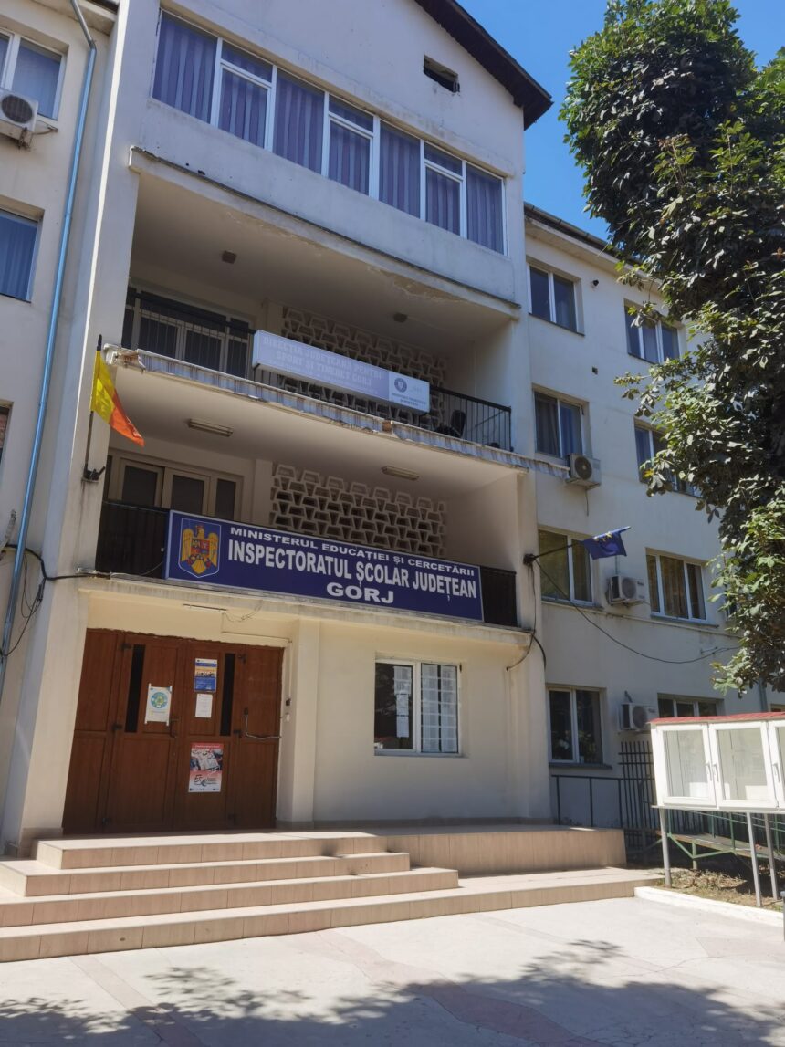 Facade of a white multi-story government building with stairs and a blue sign reading 'Inspectoratul Școlar Județean Gorj' above the entrance; Romanian flag nearby.