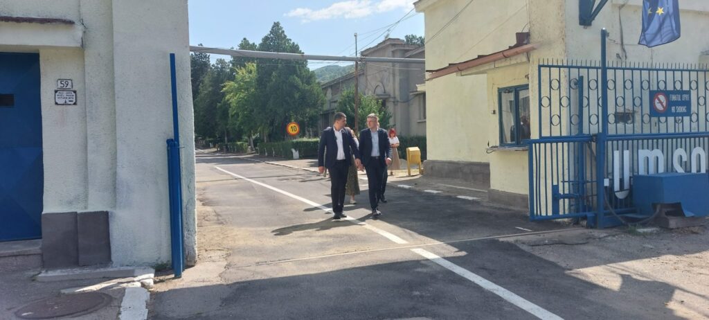 WhatsApp Image 2026-08-12 at 140111 - Jurnalul Olteniei – Știri din Craiova și Oltenia Two men in suits walk down a sunlit street between white and yellow buildings with a blue gate nearby.