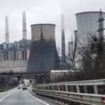 Industrial power plant with tall chimneys and cooling towers along a highway, leafless trees in the foreground on a cloudy day.