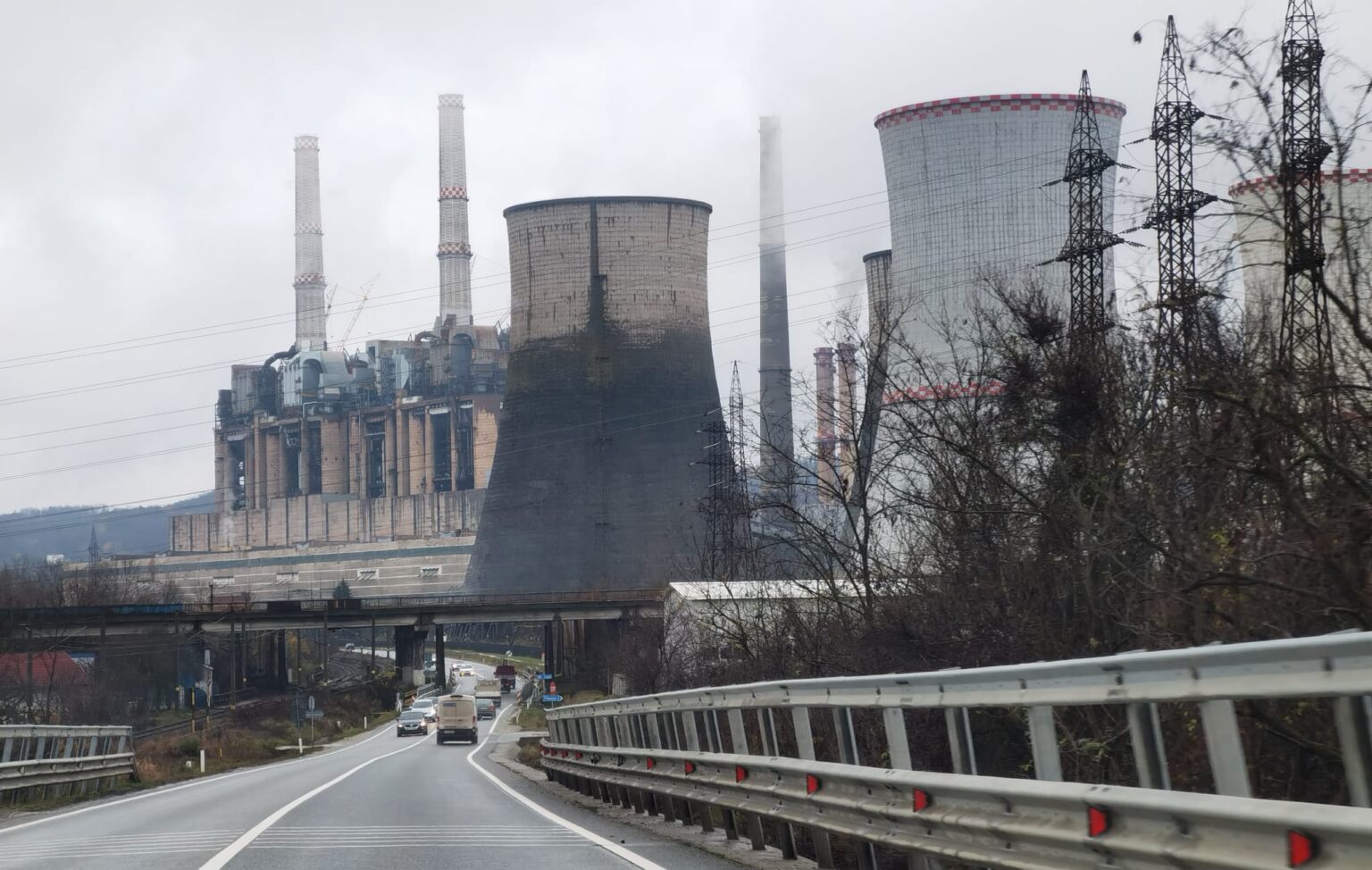 Industrial power plant with tall chimneys and cooling towers along a highway, leafless trees in the foreground on a cloudy day.
