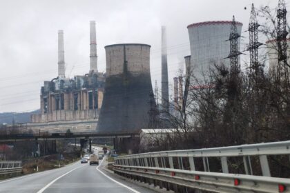 Industrial power plant with tall chimneys and cooling towers along a highway, leafless trees in the foreground on a cloudy day.