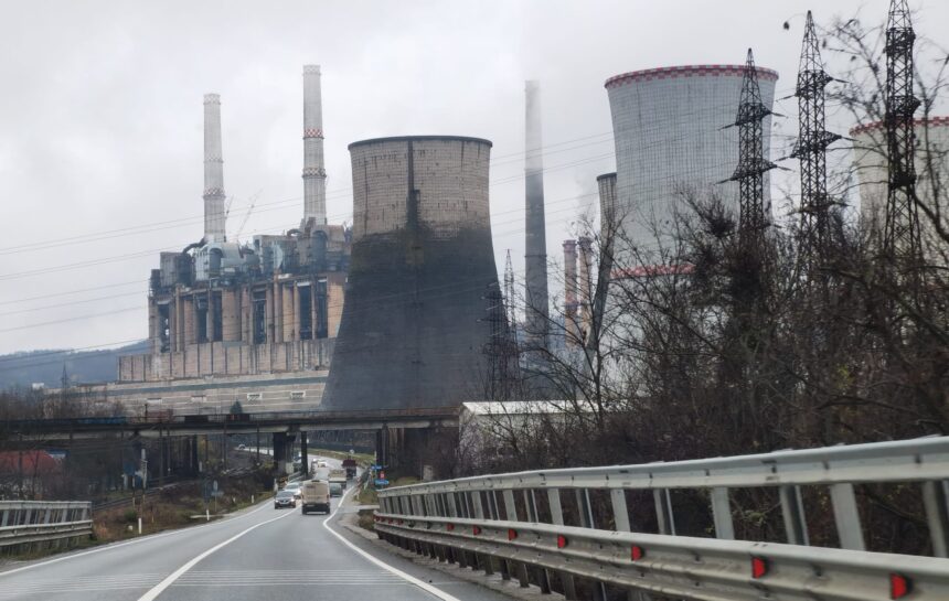 Industrial power plant with tall chimneys and cooling towers along a highway, leafless trees in the foreground on a cloudy day.