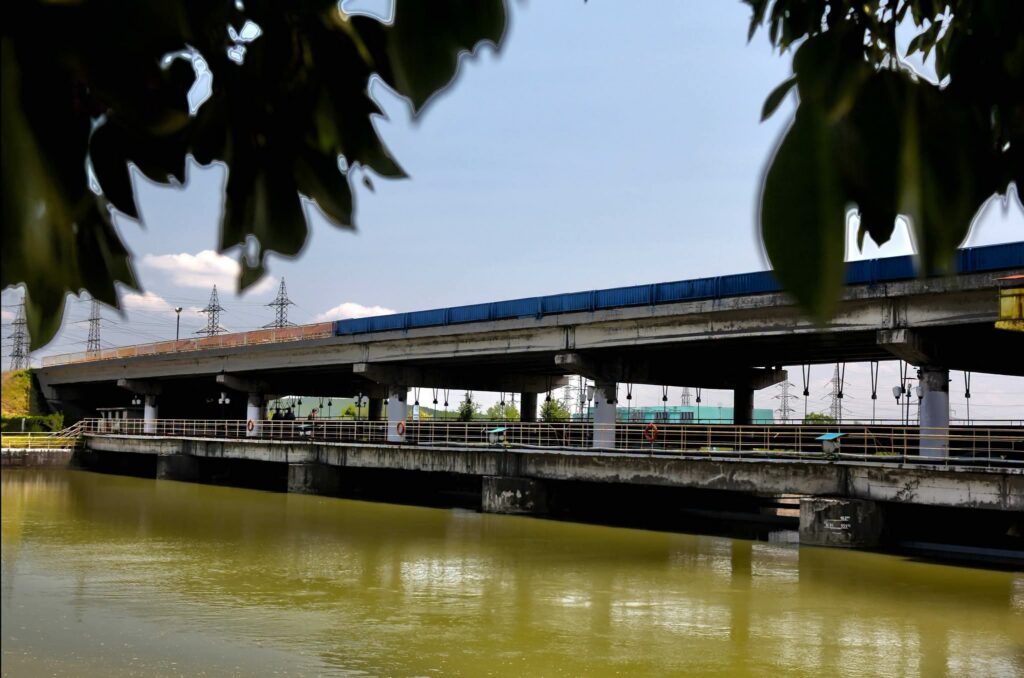 WhatsApp Image 2026-08-19 at 101846 - Jurnalul Olteniei – Știri din Craiova și Oltenia Bridge over a greenish river, seen through overhanging tree leaves in the foreground.