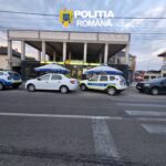 Front view of a Poliția Română building with its crest on a sign, and several police cars parked outside under blue umbrellas.