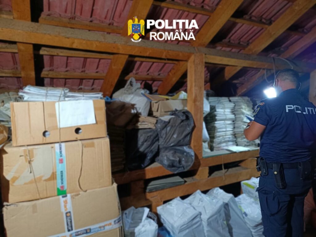 Poliția României officer in a cluttered attic filled with cardboard boxes, paper bundles, and bags.
