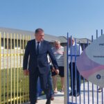 Man in a dark suit walking through a colorful gate beside a large sign reading 'Sfânta Ana' at a construction project site in Romania.