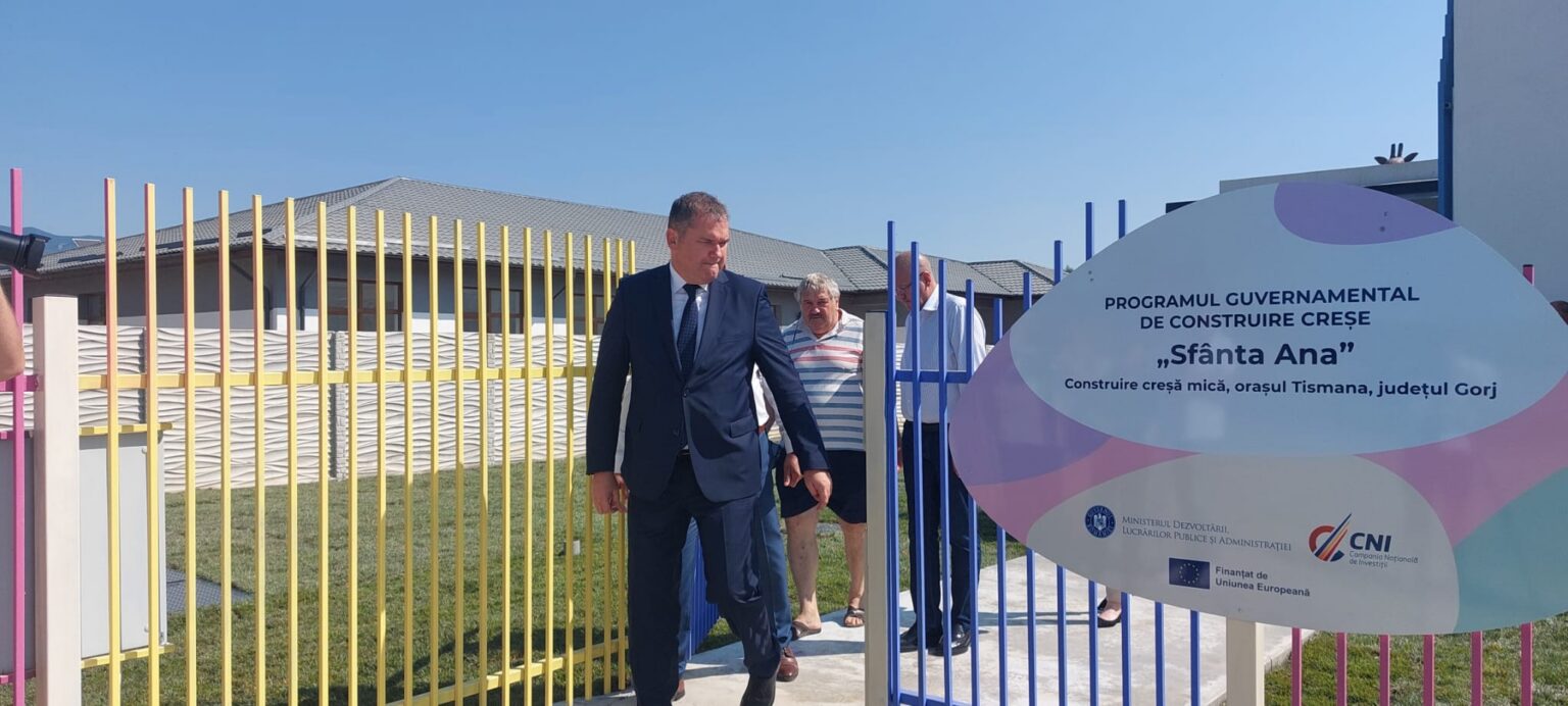 Ministrul Cseke Attila, în Gorj la inaugurarea creșei din Tismana - Jurnalul Olteniei – Știri din Craiova și Oltenia Man in a dark suit walking through a colorful gate beside a large sign reading 'Sfânta Ana' at a construction project site in Romania.