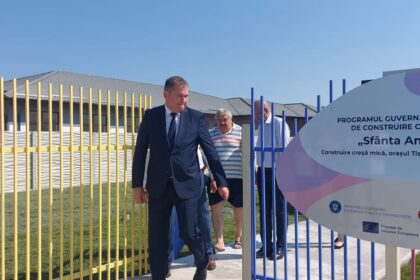 Man in a dark suit walking through a colorful gate beside a large sign reading 'Sfânta Ana' at a construction project site in Romania.