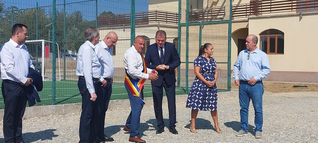 WhatsApp Image 2026-08-21 at 151217 - Jurnalul Olteniei – Știri din Craiova și Oltenia Group of seven adults in business and smart casual attire standing outside a fenced sports facility, listening to a speaker on a sunny day.