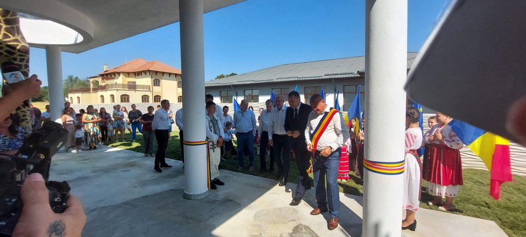 WhatsApp Image 2026-08-21 at 151218 1 - Jurnalul Olteniei – Știri din Craiova și Oltenia Outdoor ceremonial event with a man wearing a tricolor sash addressing a crowd; photographers, people in casual and traditional clothing, and blue-yellow-red flags in the background.
