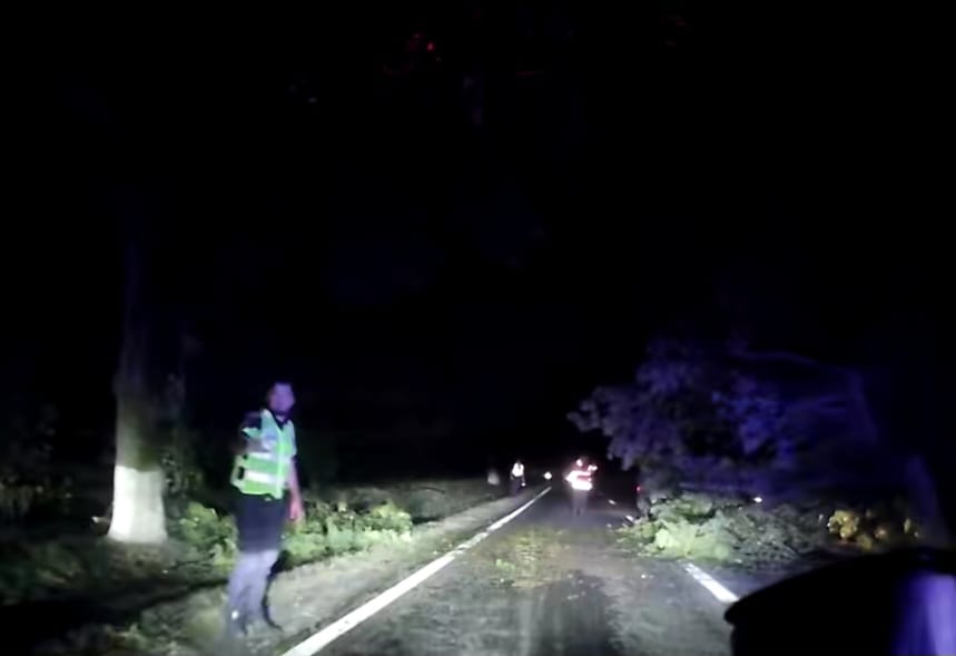 Nighttime road blocked by fallen branches; a worker in a reflective vest stands beside the lane directing traffic.