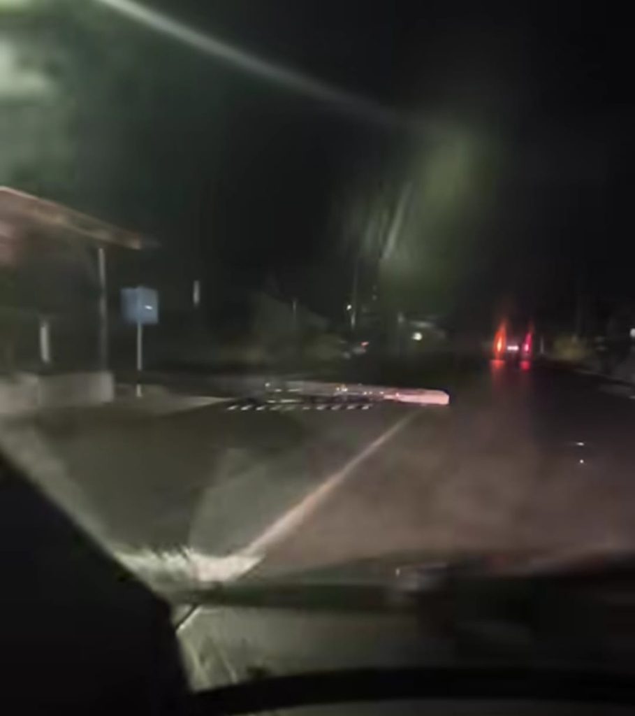 Nighttime road scene from a dashcam showing a lowered barrier across the lane ahead, street lights and dark trees in the background. A few car taillights are visible farther down the road.
