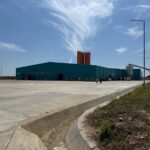 Industrial facility with a teal warehouse and tall orange silos behind, set in a wide concrete yard under a blue sky.