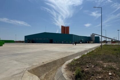 Industrial facility with a teal warehouse and tall orange silos behind, set in a wide concrete yard under a blue sky.