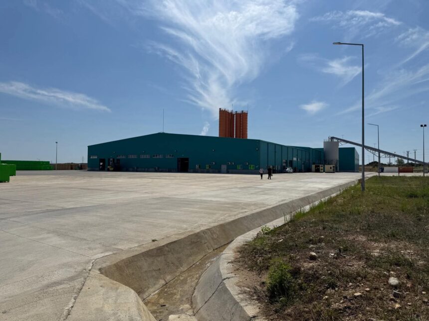 Industrial facility with a teal warehouse and tall orange silos behind, set in a wide concrete yard under a blue sky.