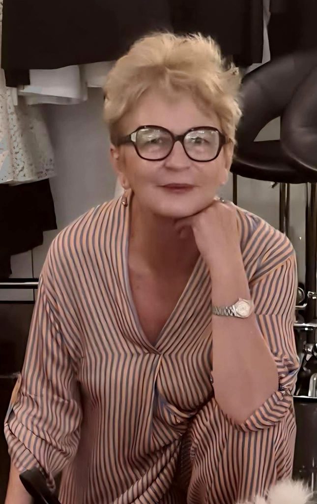 Woman with short blonde hair and glasses rests her chin on her hand, wearing a striped blouse and a silver watch.