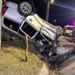 Nighttime crash scene: a silver SUV crushed against a guardrail, debris on the road, emergency crews and vehicles in the background.