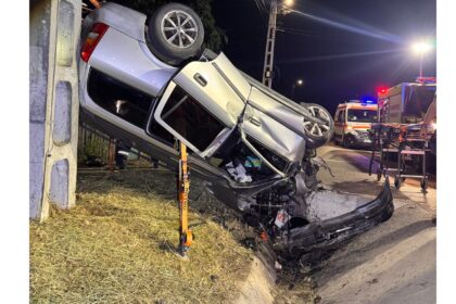 Nighttime crash scene: a silver SUV crushed against a guardrail, debris on the road, emergency crews and vehicles in the background.