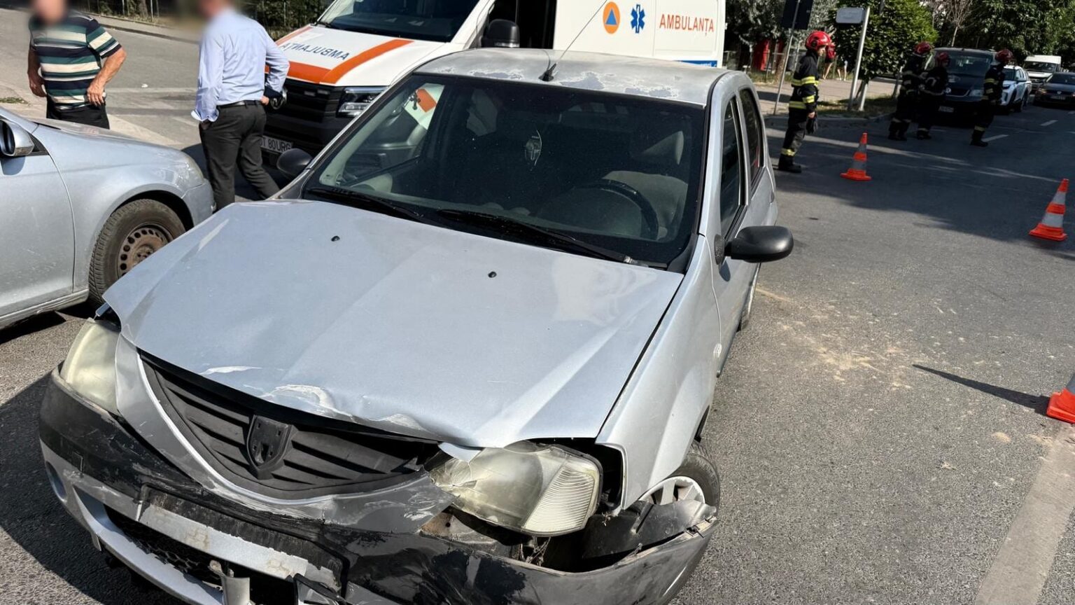 Silver sedan with severe front-end damage after a collision on a city street; an ambulance and firefighters are in the background with orange traffic cones nearby.