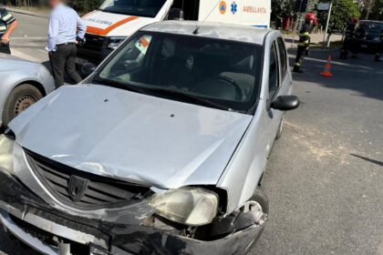 Silver sedan with severe front-end damage after a collision on a city street; an ambulance and firefighters are in the background with orange traffic cones nearby.