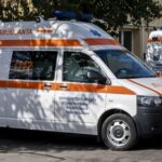 White ambulance van with orange stripe and blue Star of Life parked on a street at a sunny day edge of trees.