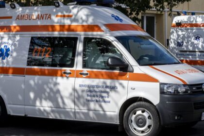 White ambulance van with orange stripe and blue Star of Life parked on a street at a sunny day edge of trees.