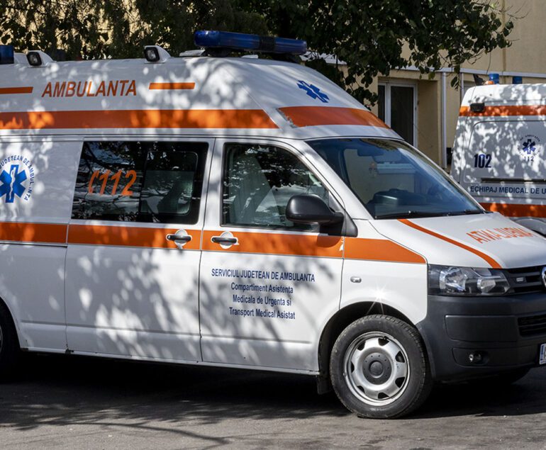 White ambulance van with orange stripe and blue Star of Life parked on a street at a sunny day edge of trees.