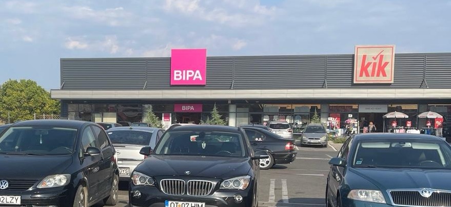 Parking lot in front of a shopping plaza with BIPA and kik storefronts and several cars parked in the foreground