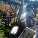 Firefighters in red helmets stabilize a blue SUV teetering over a ditch beside a guardrail as onlookers watch the rescue scene by the road.