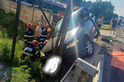 Firefighters in red helmets stabilize a blue SUV teetering over a ditch beside a guardrail as onlookers watch the rescue scene by the road.