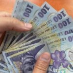 A hand holding a fan of multiple 100-euro banknotes spread out together on a wooden surface