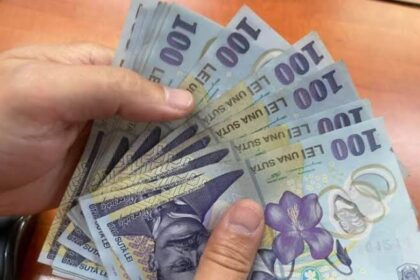 A hand holding a fan of multiple 100-euro banknotes spread out together on a wooden surface