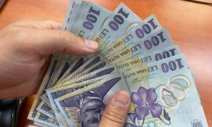 A hand holding a fan of multiple 100-euro banknotes spread out together on a wooden surface