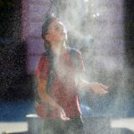 Young person in a red shirt and backpack stands with face tilted up as a fine spray of water from a fountain surrounds them.