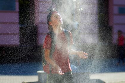 Young person in a red shirt and backpack stands with face tilted up as a fine spray of water from a fountain surrounds them.