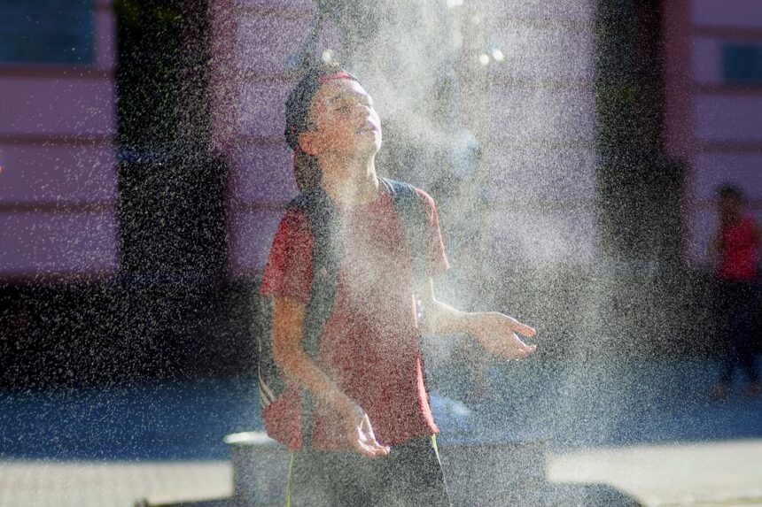 Young person in a red shirt and backpack stands with face tilted up as a fine spray of water from a fountain surrounds them.